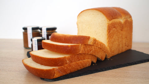 Loaf of bread with slices on a cutting board, with jars in the background.